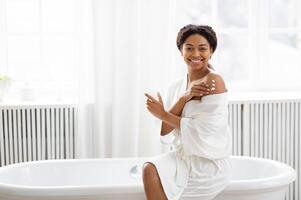A pretty millennial woman applies moisturizer on her shoulder while smiling in a bright, cozy bathroom. Soft light enhances the relaxing atmosphere of her pampering routine. photo
