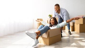 Family moving new place. Happy african american man pushing cardboard box with his wife in it, having fun while moving in new house, empty space. Moving and real estate concept. photo