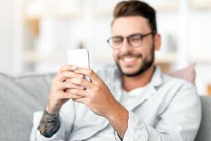 Happy millennial guy enjoying chat with lover, looking at smartphone screen and smiling, free space. Excited young man holding cellphone while resting on sofa in living room photo
