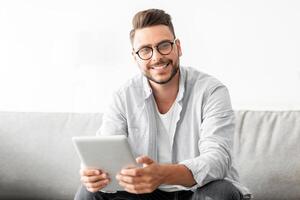 Technology concept. Excited millennial man using digital tablet, sitting on sofa at his modern apartment. Smiling guy browsing internet, resting on couch in living room, free space photo