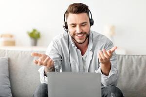 Online communication. Happy freelancer in headset having call via laptop computer, working at home, sitting on sofa. Smiling man making web conference on computer, free space photo