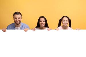 A loving family of three shares a joyful moment against a vibrant yellow backdrop, holding a blank sign. The father, mother, and daughter display warm smiles, embodying affection and connection. photo
