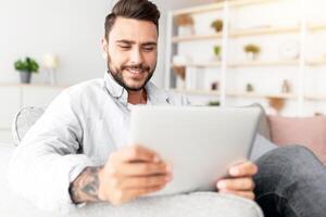 Closeup of caucasian man resting on sofa with digital tablet, home interior, free space. Happy guy sitting on couch in living room, using nice mobile app on modern pad, copy space photo