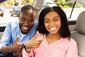 Great Perfect Car. Happy Black Couple Showing Thumbs Up Sign Gesture, Sitting In New Automobile Buying Modern Vehicle In Dealership Store Or Enjoying Vacation, Looking Out Of Window. Selective Focus photo