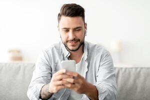 Young man with earbuds using smartphone, browsing internet and listening to music online, having call while sitting on couch at home. Cellphone application concept photo