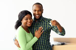 Happy african american spouses demonstrating home keys at camera and embracing, joyful black couple standing among unpacked cardboard boxes, celebrating moving in their new house, copy space photo
