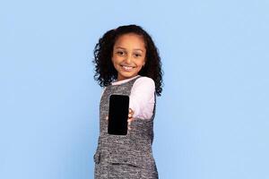 A cheerful girl with curly hair holds out a smartphone, smiling widely. She is dressed in a stylish grey outfit, surrounded by a bright blue backdrop. photo