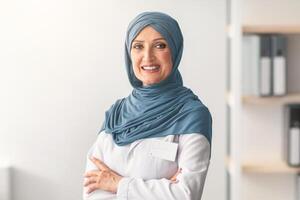 Mature Muslim Doctor Woman Wearing Hijab Smiling To Camera Standing Crossing Hands In Clinic Office. Portrait Of Professional Therapist Arab Lady Concept. Free Space For Text photo
