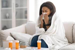 A young woman sits on a couch wrapped in a blanket, visibly unwell and sneezing into a tissue. Cold winter light shines through a nearby window, enhancing the cozy yet sick atmosphere, copy space photo