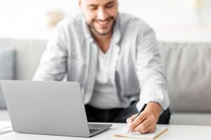 Male freelancer writing in notepad and using laptop, taking notes at workplace, using computer, noting information from internet, selective focus on male hand, closeup photo