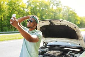 Upset black guy standing near broken car with open hood, having problem, trying to find mobile network, having no signal to call breakdown service. Road emergencies concept photo