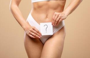 A young Caucasian woman poses confidently in white lingerie, holding a card with a question mark in front of her midsection. The background is a warm, solid color that enhances the focus on her. photo