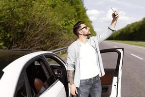 Stressed millennial Caucasian guy standing near car with cellphone, having problem, trying to find mobile network, having no signal to call breakdown service. Road emergencies, no phone connection photo