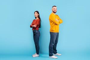 A young married couple displays a playful dynamic, standing back to back against a blue backdrop. Their casual attire and warm smiles reflect love and connection. photo