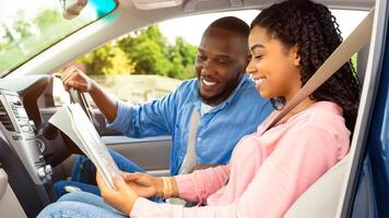 Enjoying Travel Concept. Profile side view of smiling African American couple holding and looking at paper map sitting inside luxury car, choosing best way to their trip destination, going on vacation photo