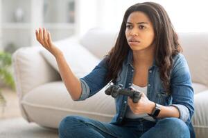 A young black woman sits on a comfortable couch in a bright living room, holding a gaming controller with a confused expression. It appears she is experiencing a challenging moment in her game. photo