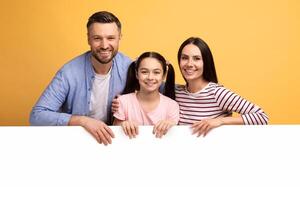 A mother, father, and their teenage daughter smile brightly while leaning on a blank white surface. The cheerful setting emphasizes their connection and warmth, showcasing a loving family bond. photo