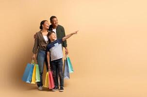 A joyful African American family stands together, holding colorful shopping bags. The teenager points excitedly at copy space, while the parents share smiles, showcasing their bond and happiness. photo
