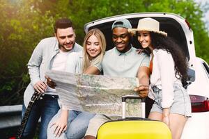 Traveling with friends concept. Happy multiracial young people sitting in open car trunk, checking map, finding their travel destination, having fun road trip adventure in summertime photo