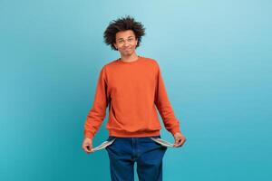 A young African American man stands confidently with a playful expression, displaying empty pockets on his blue jeans. He wears an orange sweater, contrasting with the vibrant blue backdrop. photo