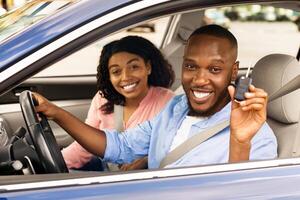 Own Car. Smiling African Amercan Couple Holding And Showing Key, Sitting In New Automobile Buying Modern Vehicle In Dealership Store, Looking Out Of Window, Happy About Luxury Purchase photo