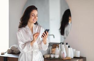 A young Caucasian woman wearing a silk robe smiles as she interacts with her phone, surrounded by skin care products in her stylish bathroom. Its a moment of relaxation and pampering at home. photo