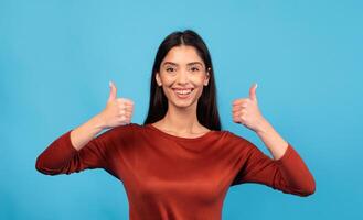 A millennial brunette woman is smiling and giving two thumbs up while standing in front of a vibrant blue background. Her joyful expression highlights a positive and confident lifestyle. photo