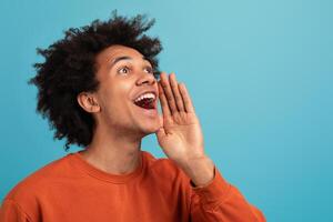 A young African American man with curly hair is joyfully shouting, showing enthusiasm. He stands isolated against a vibrant blue background, capturing a lively moment of expression, copy space photo