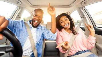 We Are Going On Vacation. Frontal view portrait of cheerful positive black guy and lady driving their automobile, dancing to music and singing favorite song, two people looking at camera photo
