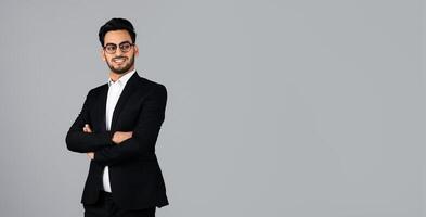 A Middle Eastern man stands confidently with crossed arms. Dressed in a sharp suit and glasses, he embodies the spirit of a millennial entrepreneur in a studio environment, copy space photo