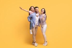 A joyful family poses together against a yellow backdrop. The father carries their daughter on his back while the mother smiles proudly. Love and togetherness are evident. photo