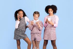 Three diverse preteen girls are joyfully engaged with their smartphones against a blue backdrop. They are experiencing childhood fun and showcasing friendship and diversity in this playful moment. photo
