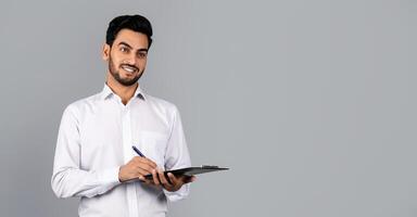 A confident Middle Eastern businessman stands in a studio environment, holding a clipboard and pen. He conveys a professional demeanor, embodying a millennial entrepreneur's lifestyle, copy space photo