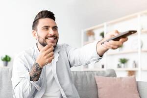 Cheerful millennial guy watching TV and eating popcorn, switching channels with remote controller while relaxing on sofa in living room interior, copy space. Free time concept photo