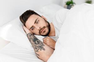 Sad millennial man lying alone in bed, suffering from loneliness and thinking of problems, copy space. Male stress and depression, sleeplessness and apathy concept. photo