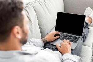 Advertisement concept. Young guy using modern laptop with blank screen, sitting on sofa, mockup. Millennial man reclining on couch and using notebook with blank display, over shoulder shot photo