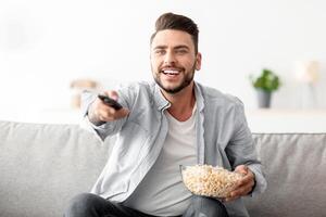 Domestic pastime. Cheerful man resting on couch switching channels with remote controller and eating popcorn, searching his favorite television show, having domestic leisure, copy space photo