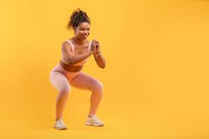 A woman wearing light pink workout attire is energetically doing squats in a vibrant yellow studio. She focuses on her form while exercising with enthusiasm and determination, copy space photo