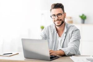 Portrait of freelancer man sitting at desk with laptop computer at home office, looking and smiling at camera, free space. Happy entrepreneur guy enjoying remote work and self-employment photo