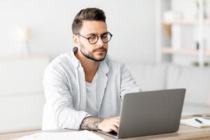 Concentrated self-entrepreneur working with laptop at home office, sitting at desk and using pc computer for remote work, copy space. Male freelancer enjoying distant job opportunities photo