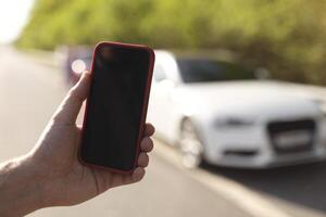 Closeup of young man showing mobile phone with empty screen for mockup, standing near his car outdoors, offering place for auto rentals or gps navigation app for summer travel photo