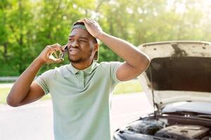 Unhappy black man standing near his broken car with open hood, experiencing engine failure, having mobile phone conversation, calling breakdown road service on smartphone photo