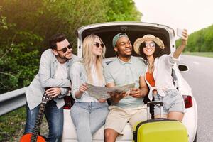 Group of best friends checking map during road trip, sitting in car trunk, taking selfie on their journey. Multiracial young people resting after long drive and having fun. Hitchhiking concept photo