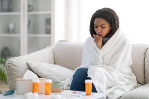 A black woman sits on a couch, wrapped in a blanket, coughing and surrounded by medicine bottles and tissues, indicating she might be feeling unwell at home, copy space photo