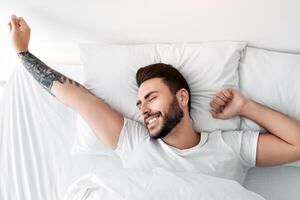 Awakening concept. Happy rested wellslept man waking up in morning and stretching hands, smiling with closed eyes while lying in comfortable bed in bedroom, top view photo
