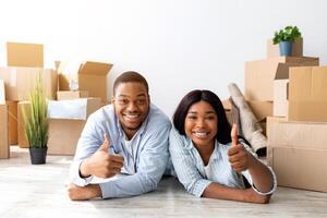 Happy black couple recommending real estate agency, showing thumbs up while lying on floor in new apartment among cardboard boxes and smiling to camera photo
