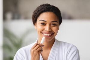 A cheerful African American woman in a bathrobe smiles while applying lip balm. The cozy atmosphere and natural light create a warm and inviting moment focused on self-care. photo