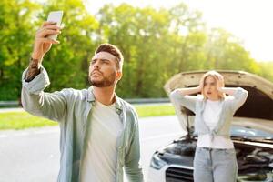 Sad young man standing on road next to his broken car together with terrified girlfriend, raising hand with mobile phone, looking for network signal, trying to call roadside assistance. No connection photo