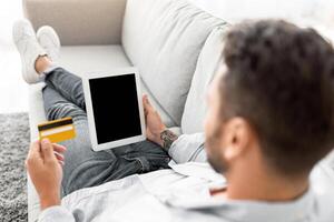Easy payments, e-commerce, online delivery concept. Young man using tablet pc and credit card, buying goods in internet store, sitting on sofa at home. Mockup for shopping app or website design photo