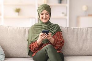 Happy Senior Middle-Eastern Woman Using Cellphone Texting And Using Mobile Application On Smartphone Sitting At Home. Mature Arab Female Browsing Internet Networking Via Phone. People And Gadgets photo
